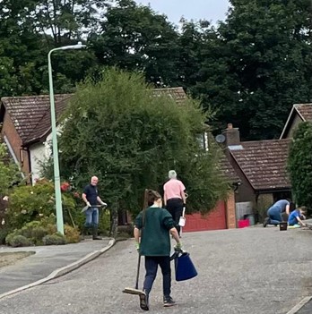 Multiple people with brooms and rubbish bags clearing up a road of weeds and moss