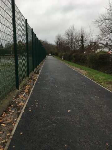 Tarmac path going into the distance between high green fencing for tennis courts and a hedgerow.